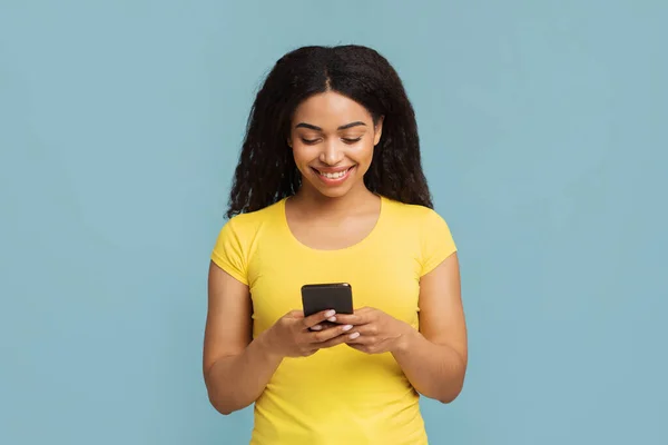 Gran aplicación. Mujer afroamericana interesada con smartphone en las manos de pie sobre fondo azul — Foto de Stock