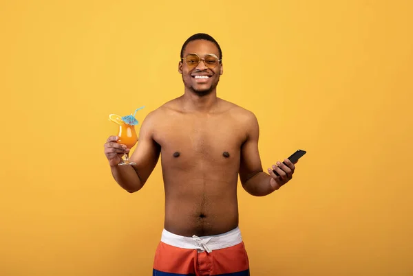 Üstsüz siyah adam akıllı telefonuyla poz veriyor ve sarı stüdyo arka planında lezzetli tropikal kokteyl. — Stok fotoğraf