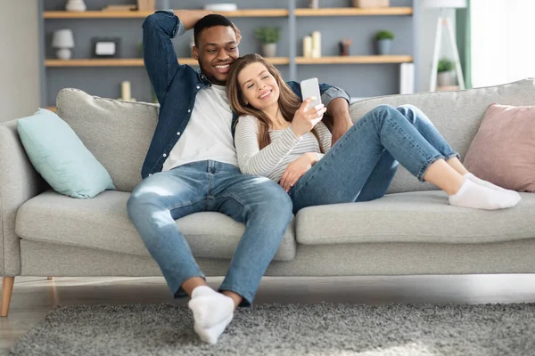 快乐的多文化配偶一起坐在沙发上，用智能手机放松一下 — 图库照片