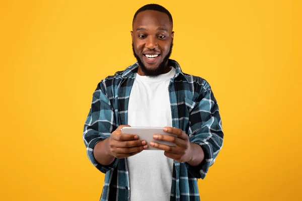 Homem Millennial preto feliz usando Smartphone jogo de jogar, fundo amarelo — Fotografia de Stock