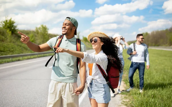 Millennial resenärer med ryggsäckar promenader längs vägen, liftar på landsbygden. Grupp människor som har autostop resa — Stockfoto