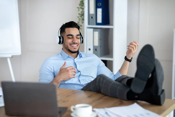 Vreugdevol Arabisch bedrijf werknemer luisteren naar muziek in hoofdtelefoon en het spelen van virtuele gitaar aan het bureau in modern kantoor — Stockfoto