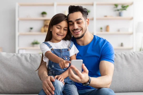 Sorrindo pai e filha sentados no sofá, usando telefone — Fotografia de Stock