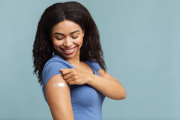 Реклама про вакцинацію проти коронавірусу. Щаслива вакцинована чорна леді демонструє руку з гіпсом на синьому фоні — стокове фото