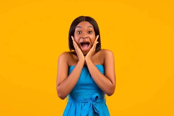 Mulher afro-americana excitada em vestido de verão gritando em emoção, de mãos dadas perto do rosto, fundo laranja — Fotografia de Stock