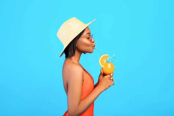 Side view of graceful black woman in swimsuit drinking cool refreshing tropical cocktail on blue studio background — Stock Photo, Image