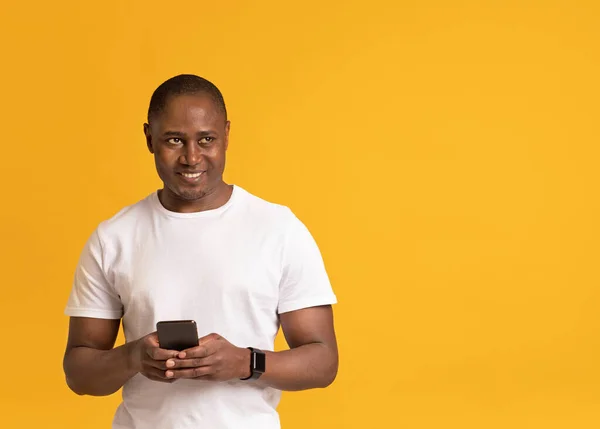 Усміхнений дорослий 40s м'язистий привабливий афроамериканський чоловік у білій футболці, що друкує на смартфоні з порожнім екраном — стокове фото