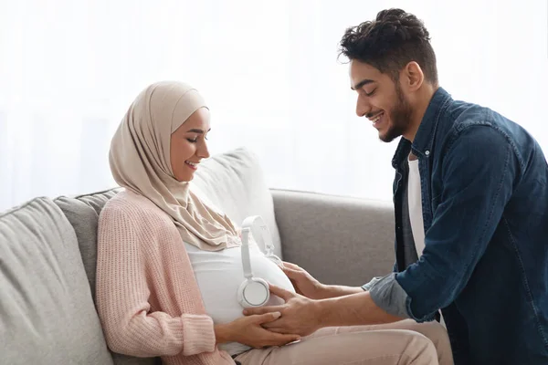 Milující arabský manžel umístění bezdrátových sluchátek na těhotné manželky břicho — Stock fotografie