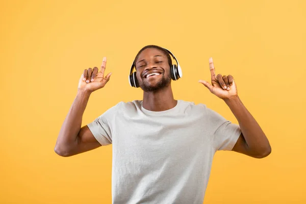 Humor juguetón. Hombre negro feliz escuchando música en auriculares, bailando y levantando los dedos sobre fondo amarillo — Foto de Stock