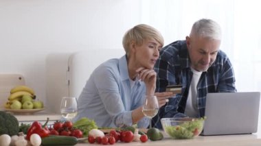 Modern mutfakta dizüstü bilgisayar ve kredi kartı kullanan orta yaşlı bir çift.