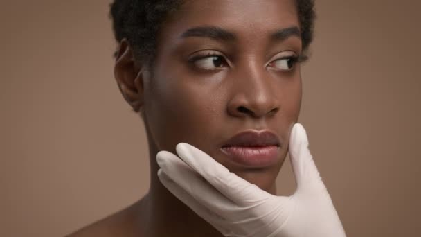 Косметический хирург касаясь афроамериканских женщин лицо, бежевый фон, обрезанный — стоковое видео