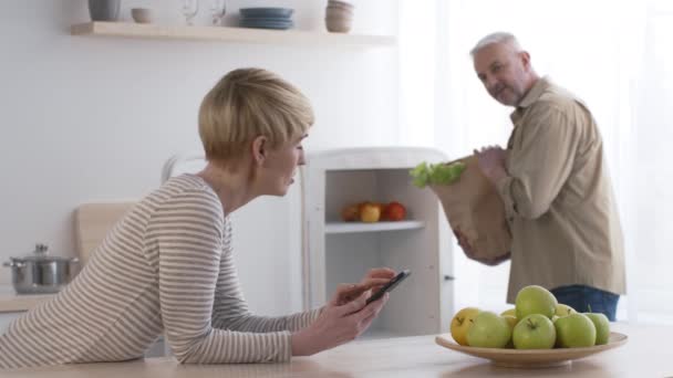 Kadın akıllı telefon kullanıyor, koca mutfaktan alışveriş yaptıktan sonra bavulunu açıyor. — Stok video