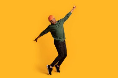 Sarı arka planda parmak uçlarında duran neşeli, Afro-Amerikalı genç adam.