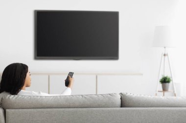 Afro-Amerikalı kadın televizyon izliyor düz ekran plazma televizyona uzaktan kumandayla işaret ediyor.