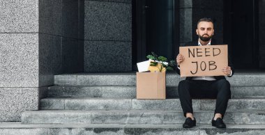 Orta yaşlı iş adamı iş merkezinin yanında oturuyor. Yanında bir kutu ofis malzemesi ve işe ihtiyacı olan bir poster var.