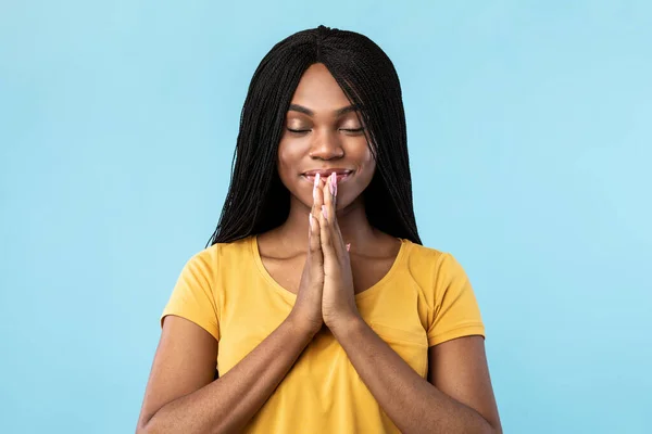 Feliz senhora afro-americana rezando com os olhos fechados, fundo azul — Fotografia de Stock