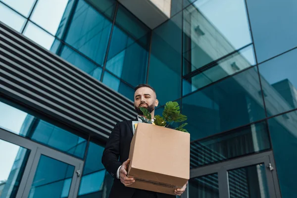 Concepto de desempleo. Empresario despedido caminando al aire libre cerca del edificio de oficinas con caja de cosas personales, espacio libre — Foto de Stock