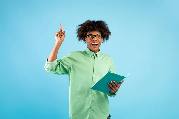 Feliz estudiante afroamericano masculino apuntando con el dedo hacia arriba, teniendo idea creativa, sosteniendo el cuaderno en el fondo azul del estudio — Foto de Stock