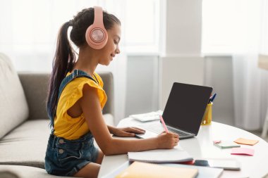 Masada oturan, dizüstü bilgisayar kullanan, ders kitabı yazan bir kız.