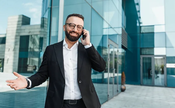 Feliz hombre de negocios exitoso hablando en el teléfono inteligente, de pie al aire libre contra el centro de oficina moderno, espacio libre — Foto de Stock