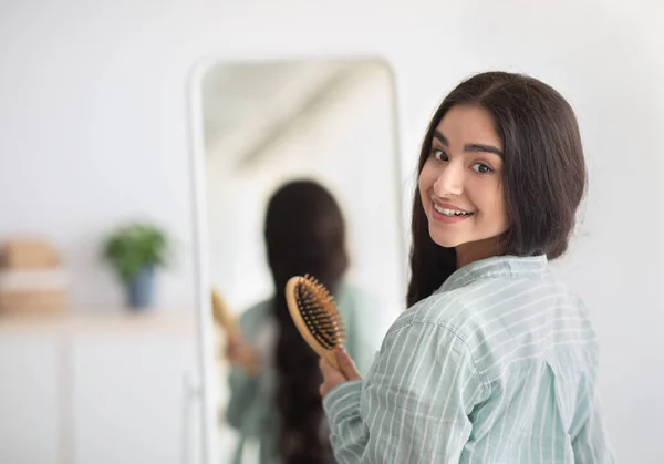 Wanita India yang menawan dengan rambut indah panjang berdiri dengan sikat kayu di depan cermin, melihat ke dalam ruangan kamera — Stok Foto