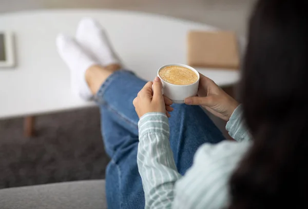 Onherkenbare Indiase vrouw zit op de bank met een kopje schuimige cappuccino, zet haar voeten op salontafel thuis — Stockfoto