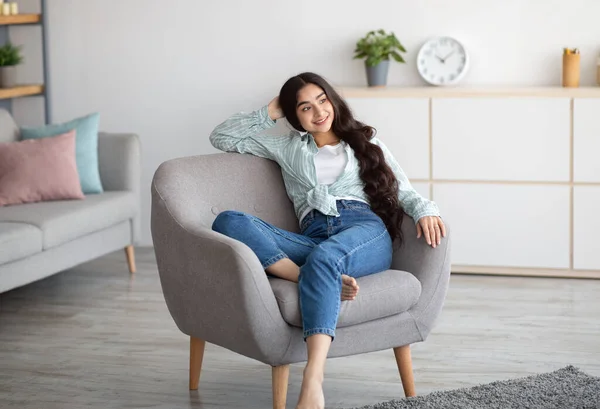 Gorgeous Indian woman sitting in comfy armchair, enjoying lazy morning, having peaceful weekend at home