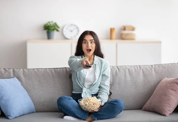 Toute la longueur de femme indienne choquée assis sur le canapé, manger du pop-corn, changer de chaînes, regarder la télévision à la maison — Photo