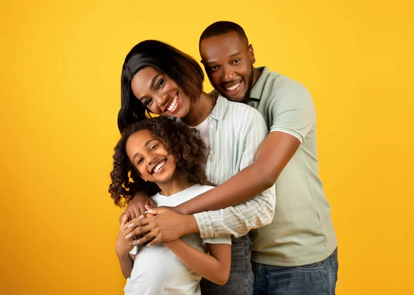 Retrato de família negra feliz de três com linda filha abraçando e sorrindo para a câmera sobre fundo amarelo — Fotografia de Stock