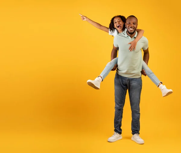 Alegre afro-americano pai dando piggyback passeio para filha, menina apontando para o espaço livre no fundo amarelo — Fotografia de Stock