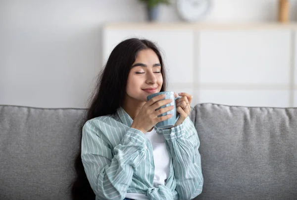 Портрет красивой индийской женщины, отдыхающей на диване, нюхающей ароматный кофе с закрытыми глазами у себя дома — стоковое фото