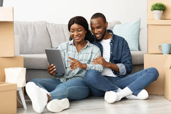 Cônjuges pretos com tablet digital sentado entre caixas de papelão em novo plano — Fotografia de Stock