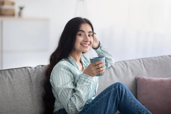 Portret van een jonge Indiase vrouw met koffiepauze, zittend op de bank met een kopje warme drank thuis — Stockfoto