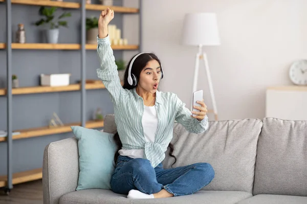 Mujer india milenaria en auriculares mirando el teléfono inteligente, levantando la mano, haciendo un gesto de victoria en casa — Foto de Stock