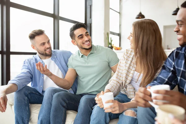 Grupo internacional de amigos com bebidas quentes conversando, sentado no sofá, tendo festa em casa — Fotografia de Stock
