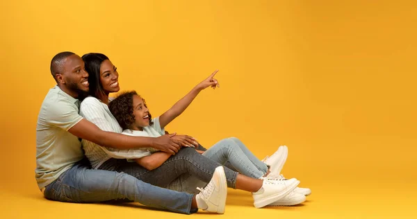 Alegre familia afroamericana sentada en el suelo, linda chica apuntando con el dedo al espacio de copia sobre fondo amarillo, panorama — Foto de Stock