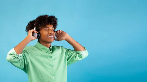 Африканський американець, який любить музику з заплющеними очима в бездротових навушниках, стоїть на синьому фоні, панорама — стокове фото