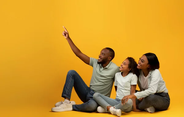 Mira aquí. Hombre negro feliz mostrando algo a su familia, sentado sobre un fondo amarillo, señalando con el dedo el espacio de copia —  Fotos de Stock