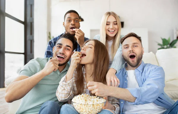 Diversos jovens amigos comendo pipocas enquanto assistem TV, se divertindo juntos em casa — Fotografia de Stock