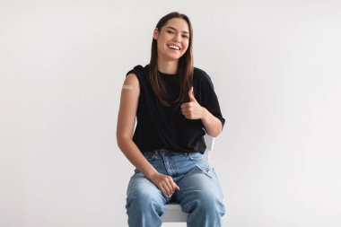 Antiviral enjeksiyondan sonra kolları aşılanmış mutlu genç bir bayan ışık stüdyosunun arka planında baş parmak hareketlerini gösteriyor.