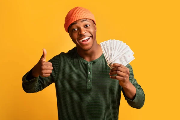 Большая прибыль. Веселый черный человек держит доллар наличными и показывает большой палец вверх — стоковое фото