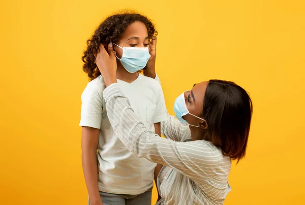 Prevención del Coronavirus. negro madre poniendo en protección quirúrgica máscara en hijas cara, fondo amarillo —  Fotos de Stock
