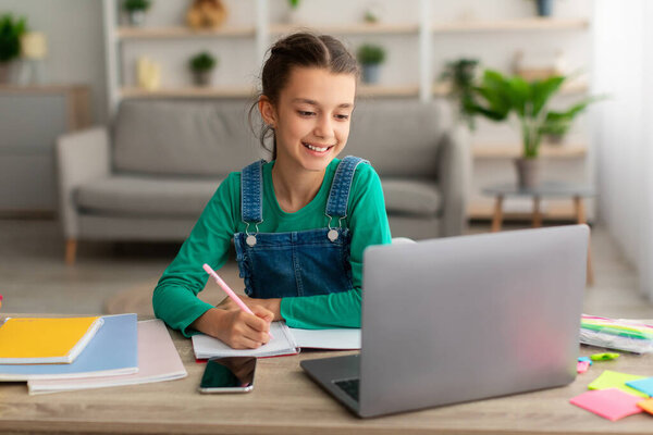 Girl sitting at table, using laptop, writing in notebook