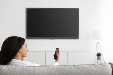 Akşamları eğlence. Rahat siyahi bayan televizyon seyrediyor. Uzaktan kumandayı siyah ekranlı düz ekrana doğrultuyor.