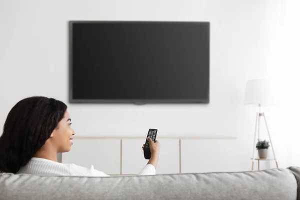 Plezier in de avond. Ontspannen zwarte dame kijken TV wijzend afstandsbediening op flatscreen televisie met zwart scherm — Stockfoto