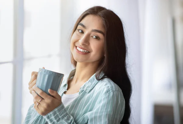 Portret van een mooie Indiaanse dame bij het raam met een kop warme drank, glimlachend naar de camera, binnen — Stockfoto