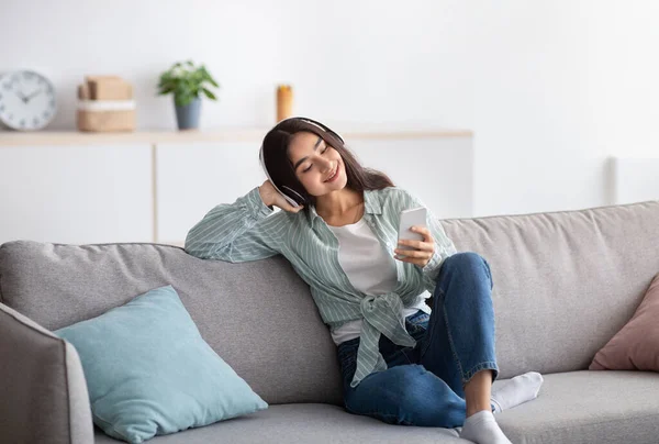 Радостная индийская леди в наушниках сидит на диване и слушает музыку на смартфоне дома, в полный рост — стоковое фото