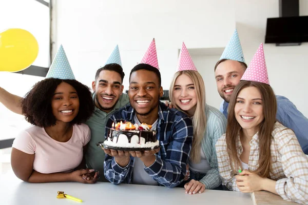 Ritratto di felici amici multietnici che indossano cappelli da festa, in posa con torta di compleanno, sorridendo alla macchina fotografica all'interno — Foto Stock