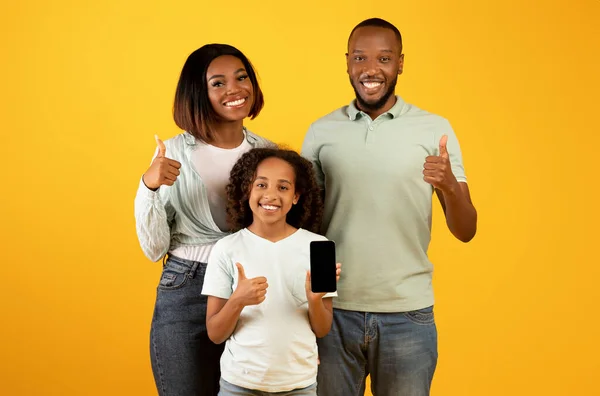 Familia afroamericana positiva de tres teléfonos inteligentes demostrando y mostrando los pulgares hacia arriba sobre el fondo amarillo —  Fotos de Stock