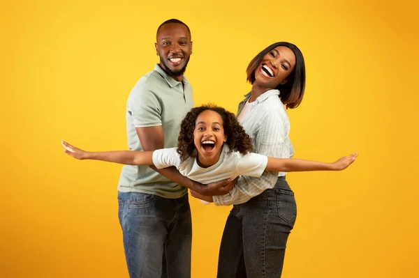 Conceito de ligação familiar. Joyful homem negro e mulher segurando filha a mãos, se divertindo sobre a parede do estúdio amarelo — Fotografia de Stock